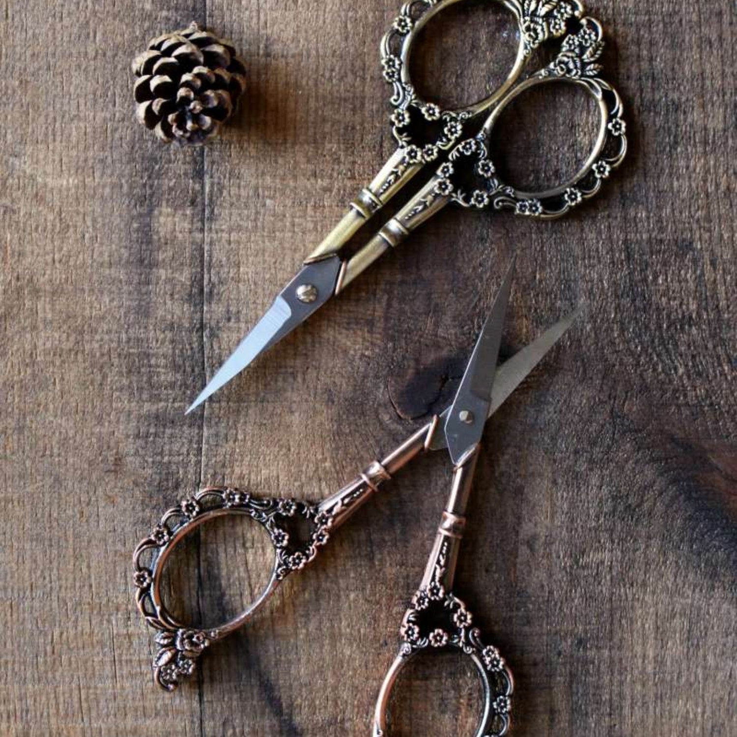 Pair of ornate scissors on a wooden surface with a pine cone.